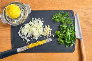 Zwiebeln und Knoblauch würfeln, Petersilie und Dill waschen, trocknen und Blätter abzupfen, Zitronenzeste abreiben, Zitrone auspressen