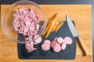 Fleischwurst pellen, in Scheiben und dann in Streifen schneiden